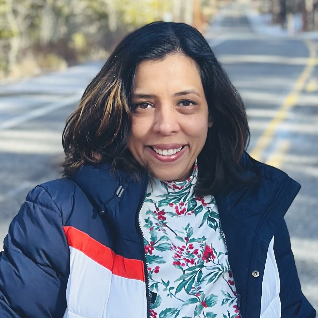 siri A woman with shoulder-length dark hair smiles outdoors, wearing a navy and white puffer jacket with a red stripe and a floral turtleneck top. The background shows a sunlit, empty road and trees.