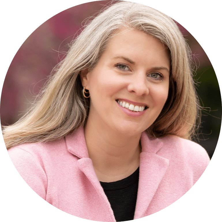 Emily Behnke-modified A woman with long, straight, grayish-blonde hair smiles at the camera. She is wearing a pink blazer over a black top, with a blurred outdoor background.