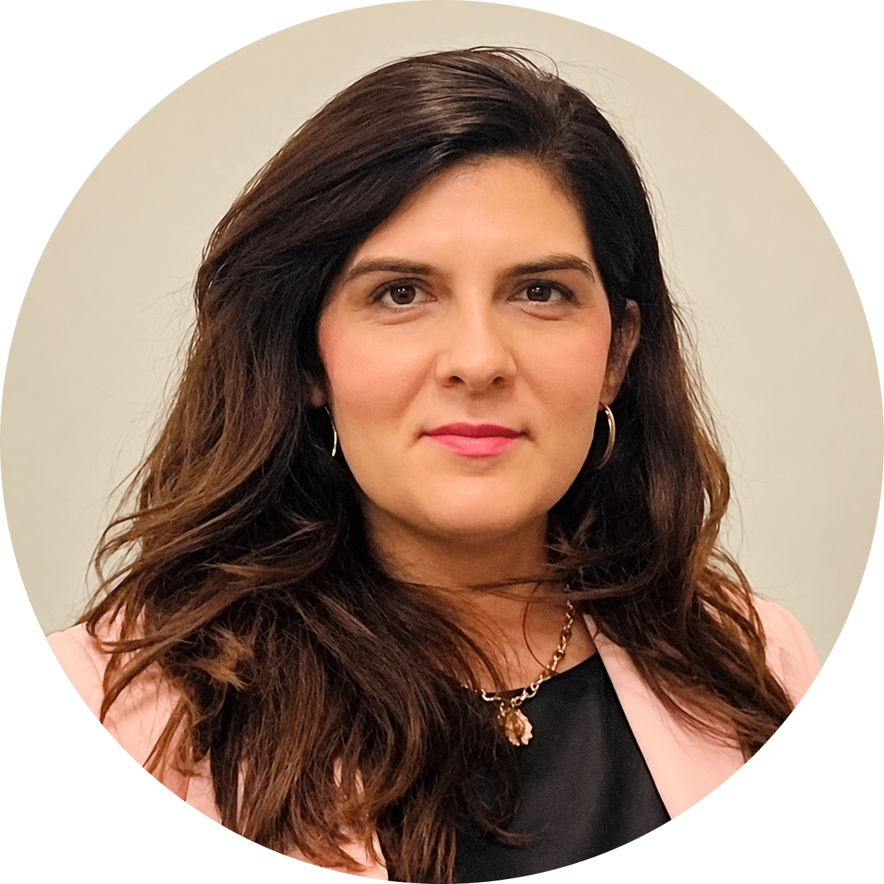 Andrada-modified Woman with long brown hair wearing a light pink blazer, black top, gold necklace, and hoop earrings, posing in front of a neutral background.