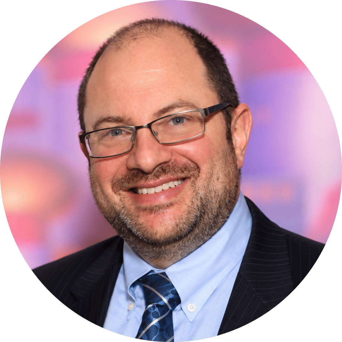 Eric A Joseph-modified A smiling man with glasses, short brown hair, and a beard, wearing a dark suit, light blue shirt, and patterned tie. The background is blurred with pink and purple tones.