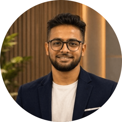 Karan Patel Headshot-modified A man with short dark hair, a beard, and glasses smiles at the camera. He is wearing a navy blazer over a white shirt. The background features a plant and wooden paneling.