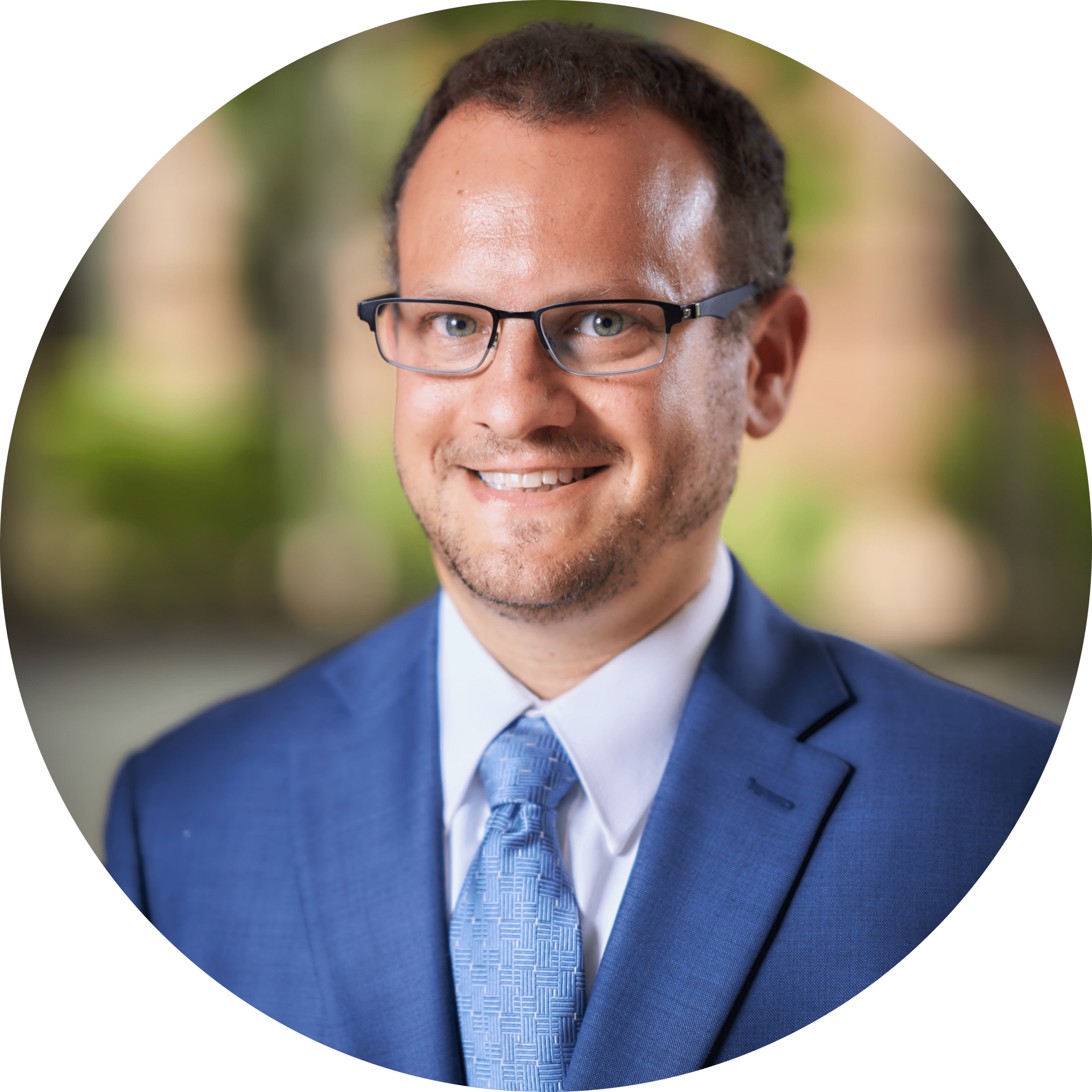 Matt Gruber Headshot-modified Smiling man with glasses in a blue suit and tie, posing in front of a blurred outdoor background.