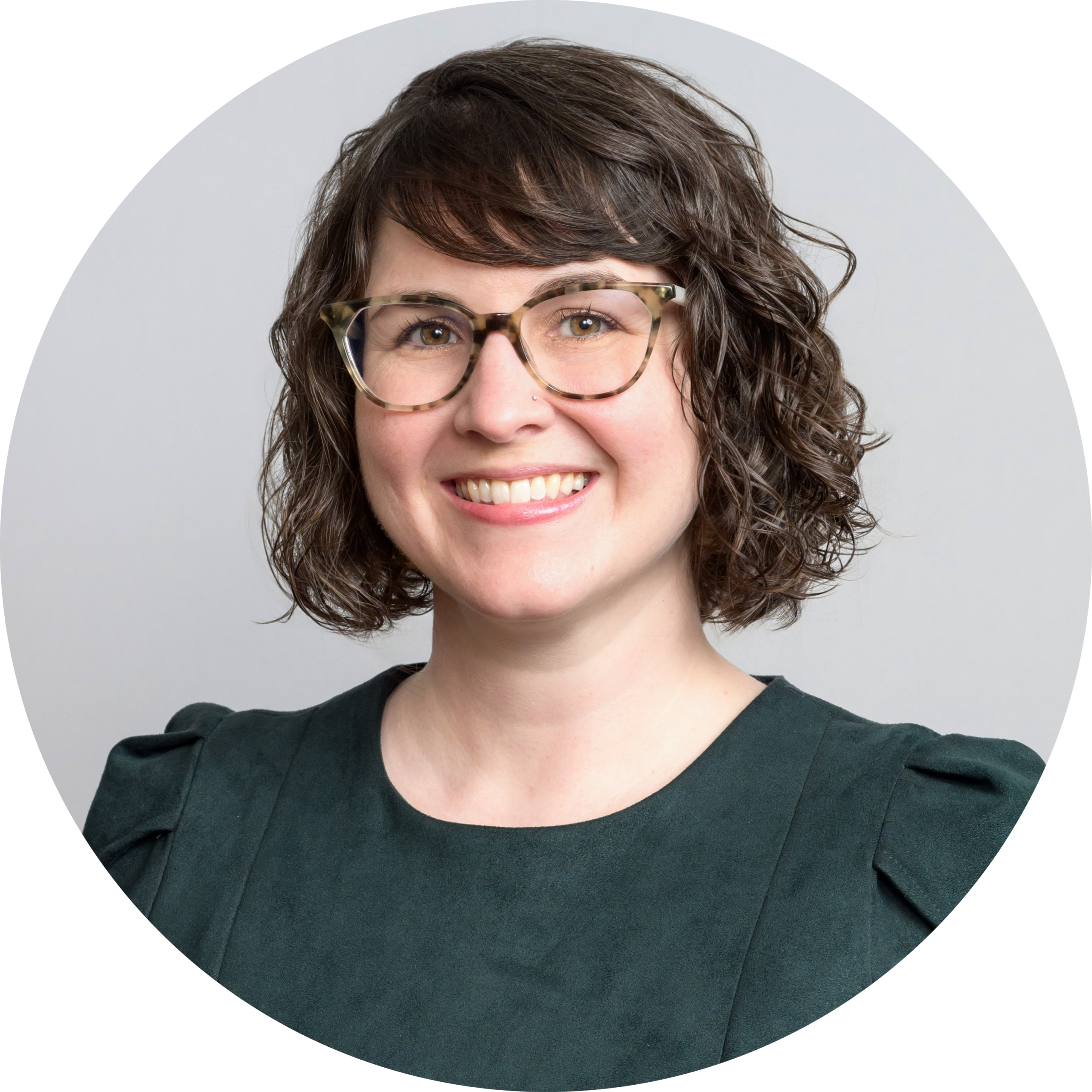 Mclear_Christine-modified Woman with short, curly brown hair wearing glasses and a dark green top, smiling at the camera against a light gray background.