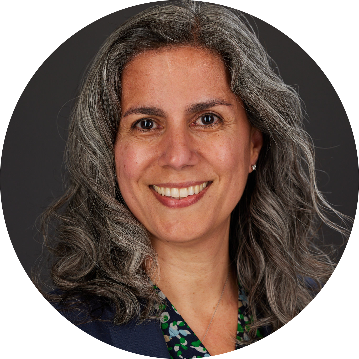 Tara McCaughey headshot-modified Smiling woman with long wavy gray hair, wearing a dark blazer and a patterned top, posing in front of a dark background.